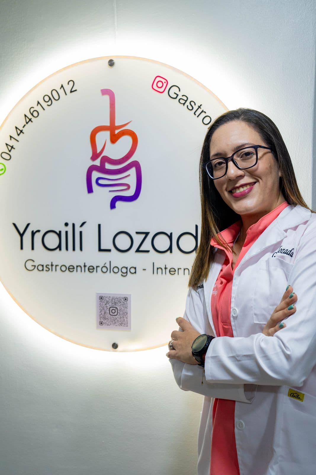 La Dra. Yraili Lozada con bata blanca y los brazos cruzados, sonriendo junto al letrero iluminado de su consultorio